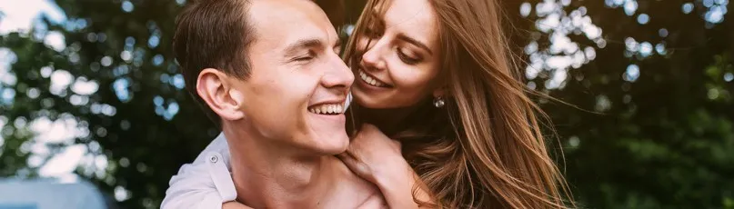 Casal sorridente ao ar livre, com a mulher a abraçar o homem por trás — Consultas