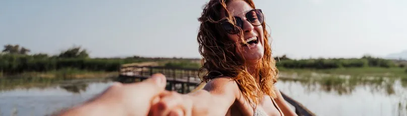 Mulher a sorrir com óculos de sol, a segurar a mão de alguém, ao ar livre junto à água — Consultas