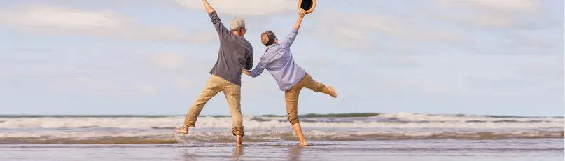 Um casal idoso a brincar na praia, com os braços levantados e uma perna levantada — Consultas