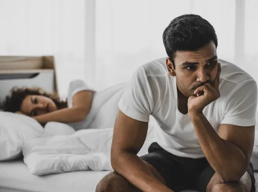 Homem sentado na cama com expressão preocupada, mulher a dormir ao fundo — Impotência