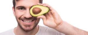 Homem sorridente segura meio abacate sobre o olho direito — 10 alimentos que melhoram a saúde e a potência