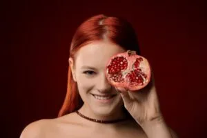 Mulher de cabelo vermelho segura uma romã partida à frente do rosto — 10 alimentos que melhoram a saúde e a potência