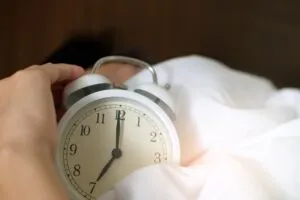 Mão a apagar um despertador branco sobre lençóis brancos — Diz-me como dormes, dir-te-ei quem és!