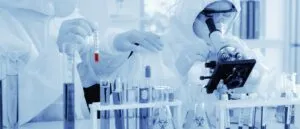 Cientistas em trajes de proteção analisam amostras com microscópio e tubos de ensaio