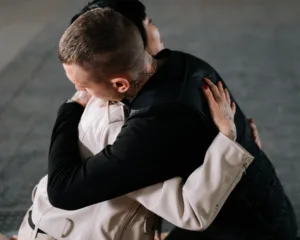 Duas pessoas abraçadas de costas, uma com tatuagens no pescoço e a outra com unha vermelha